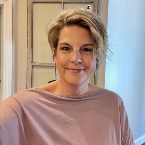 Smiling woman with short blonde hair wearing a pink top, standing indoors near a door frame.