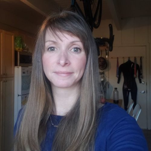 Portrait of a woman with long light brown hair wearing a blue top, indoors with a bicycle hanging in the background.